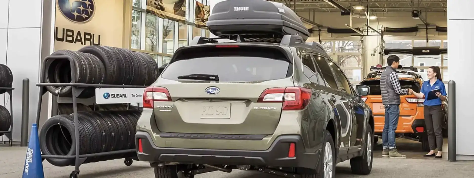 A Subaru Outback with a Thule roof cargo box sits in a Subaru service center next to a tire rack