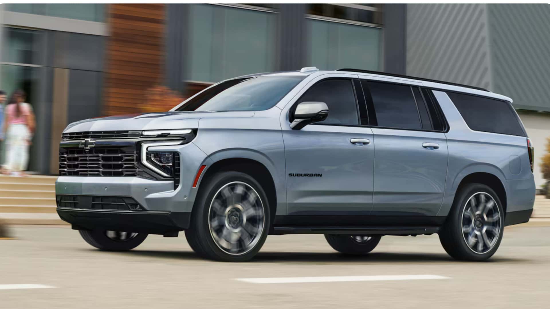 2026 Chevrolet Suburban silver SUV driving past modern city buildings in side profile action view.