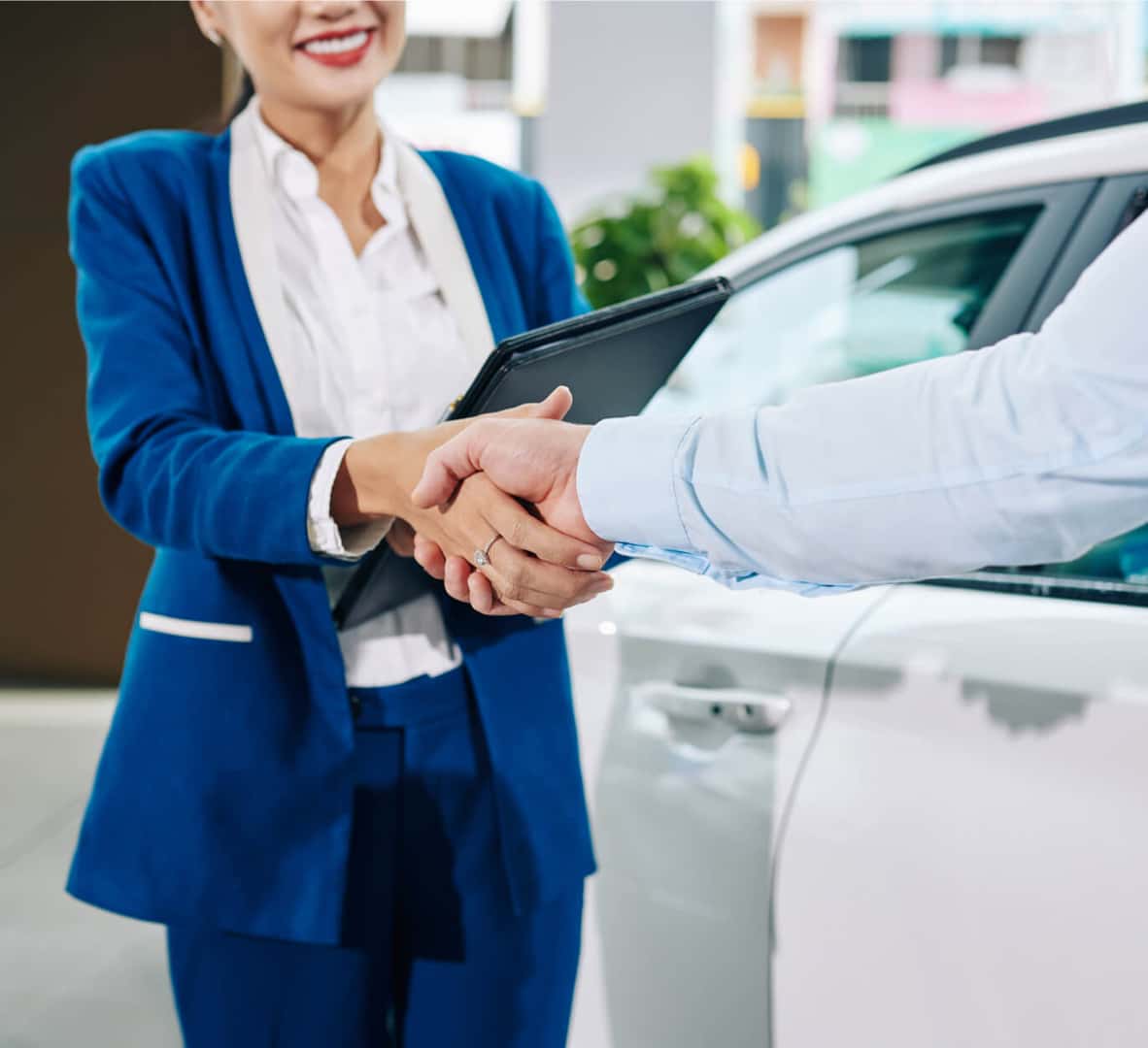 Close-up of a salesperson in a blue suit shaking hands with a customer next to a white vehicle at a dealership.