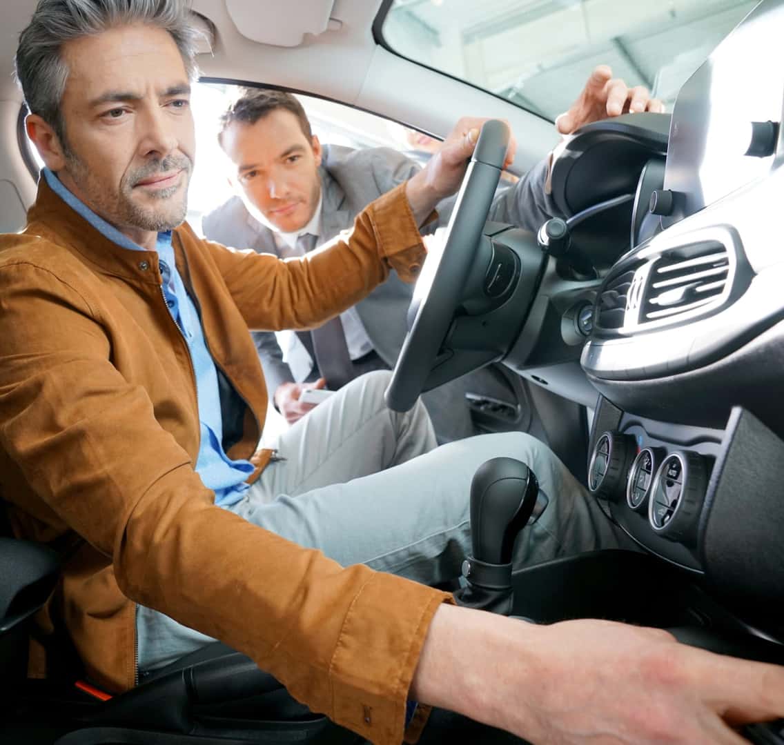 Two men are examining the interior of a car. The man in the driver's seat adjusts controls, while the other leans in attentively.
