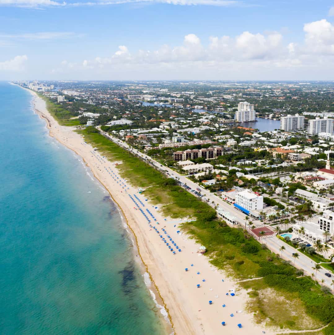 Aerial view of Delray Beach Florida featuring a long sandy beach lined with blue umbrellas beside turquoise ocean water.
