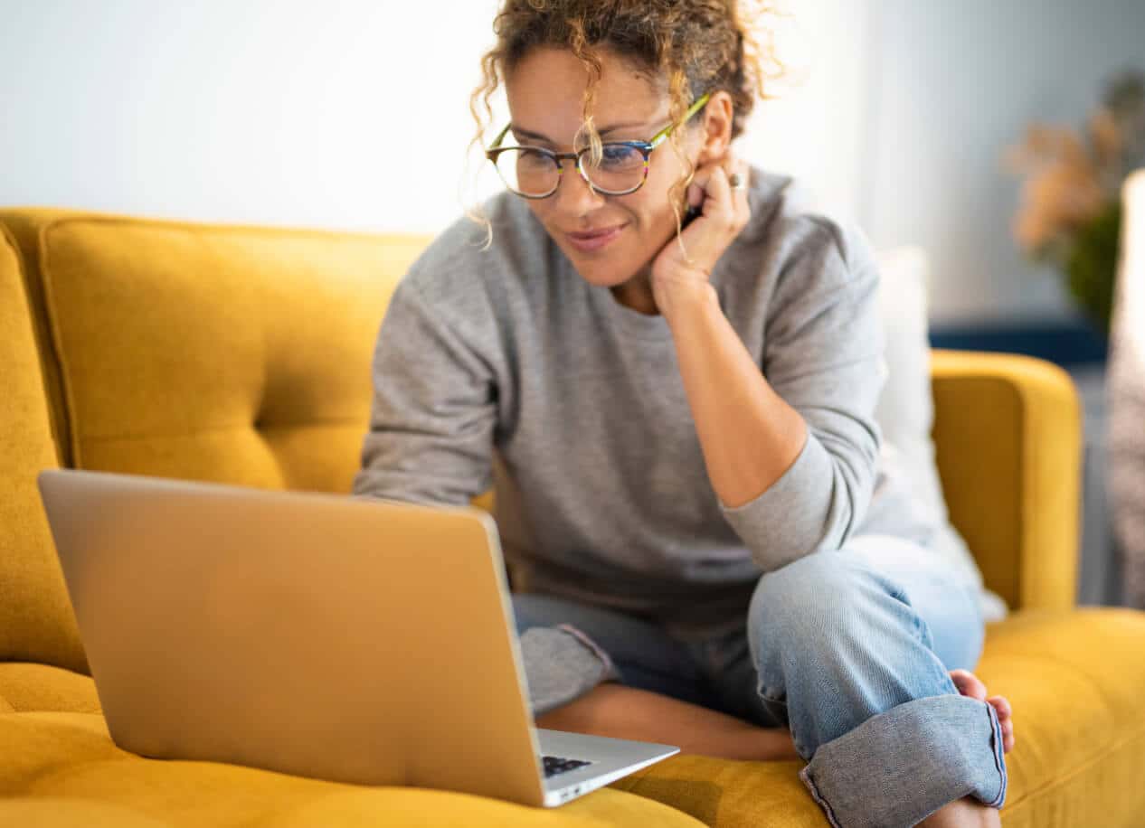 A smiling woman sits on a yellow couch in a bright room interacting with her laptop.