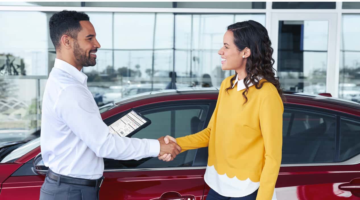 A side view of a Nissan Service Center Representative shaking hands with a woman near a red Nissan car.