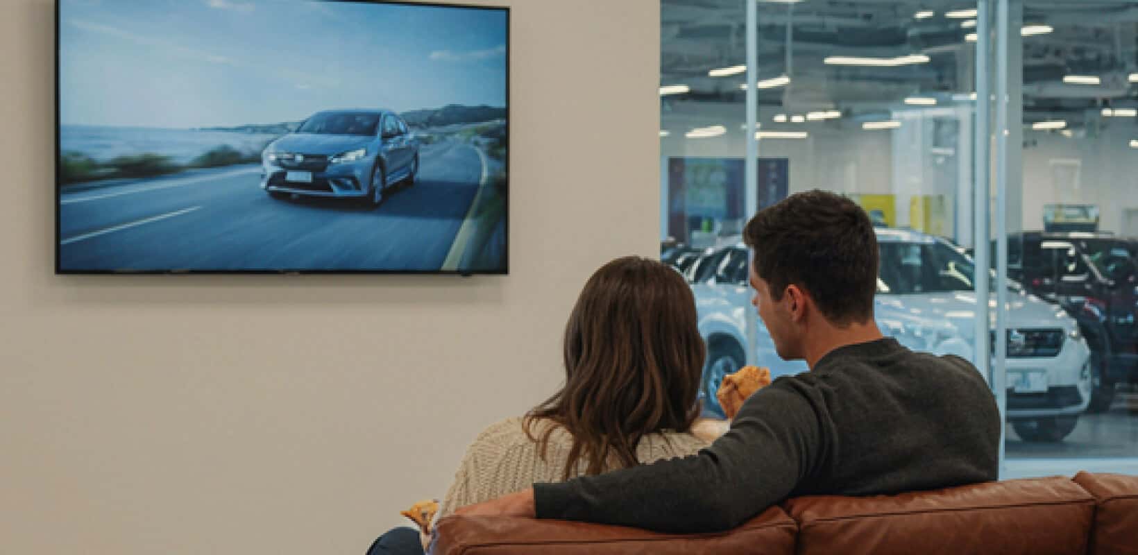 Couple on a sofa watching a TV displaying a driving car in a car dealership waiting area.