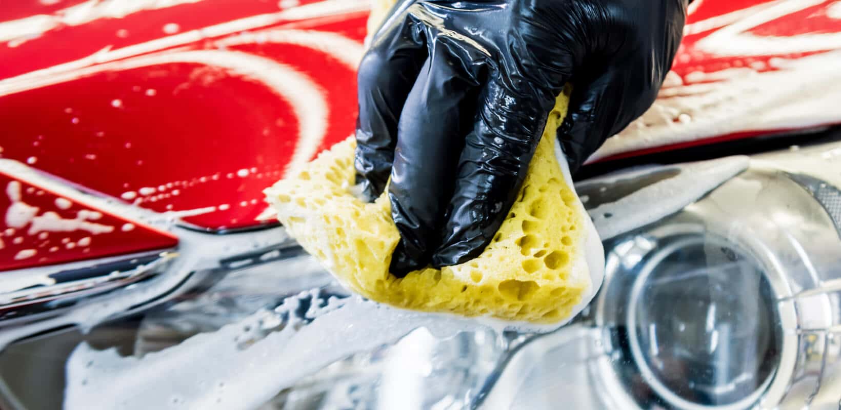 Close-up of a gloved hand washing a red car hood and headlight with a soapy yellow sponge.