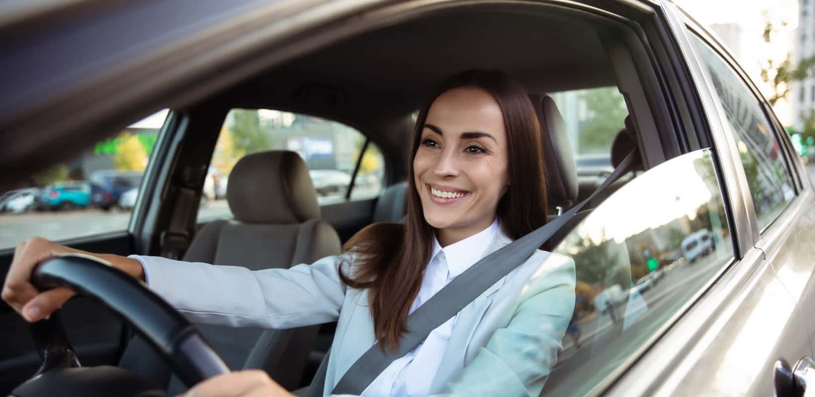 Interior view of a smiling woman driving a silver loaner sedan on a city street.
