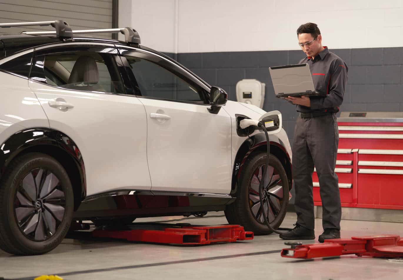 Side profile of a white Nissan Ariya EV charging while a technician diagnoses it on a laptop in a bay.