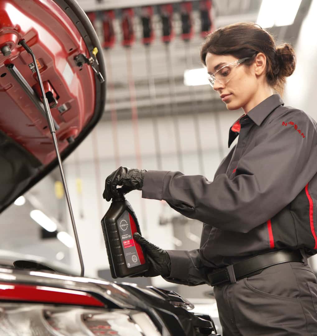 Female technician holding a bottle of Nissan motor oil under the open hood of a red car in a service bay.