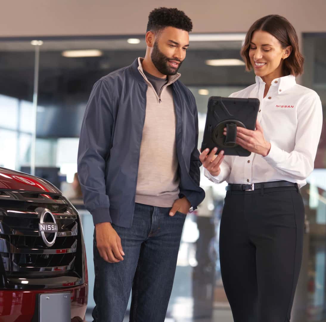 Nissan service rep showing a tablet to a customer next to a red Nissan in a dealership showroom.