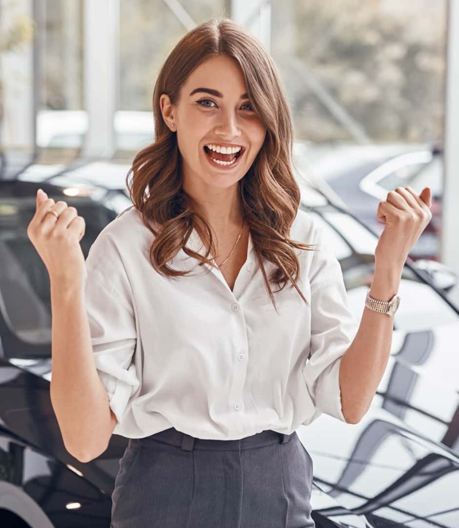 Woman celebrates a dealership award win, smiling with raised fists beside vehicles in a bright showroom.