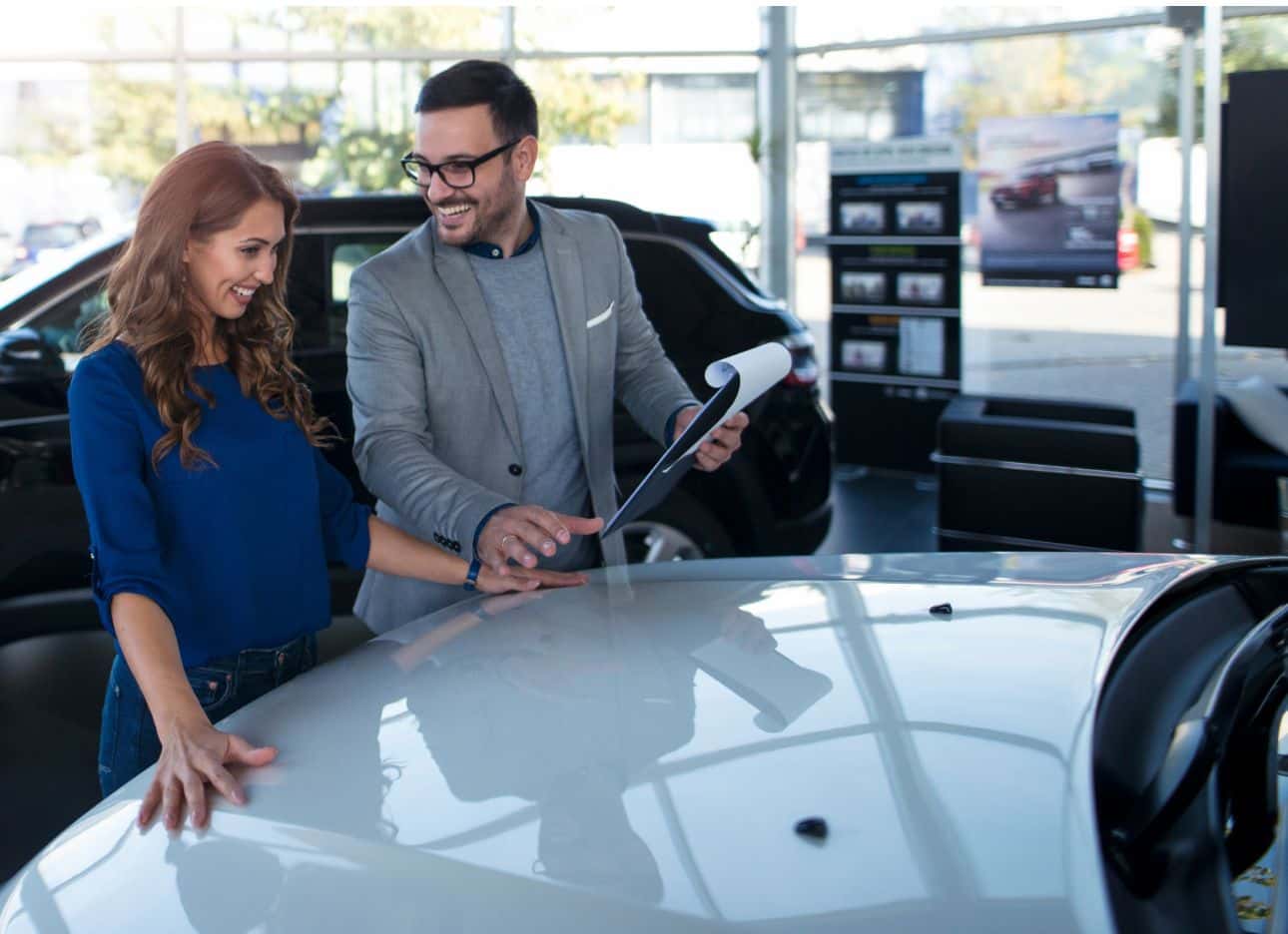 Woman browsing a vehicle with a salesperson holding paperwork inside a car dealership.