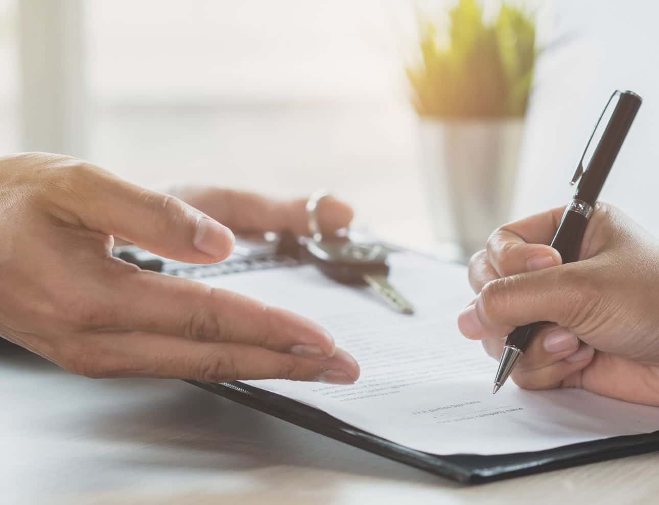 Close-up of a person signing paperwork while another person gestures beside a set of car keys.