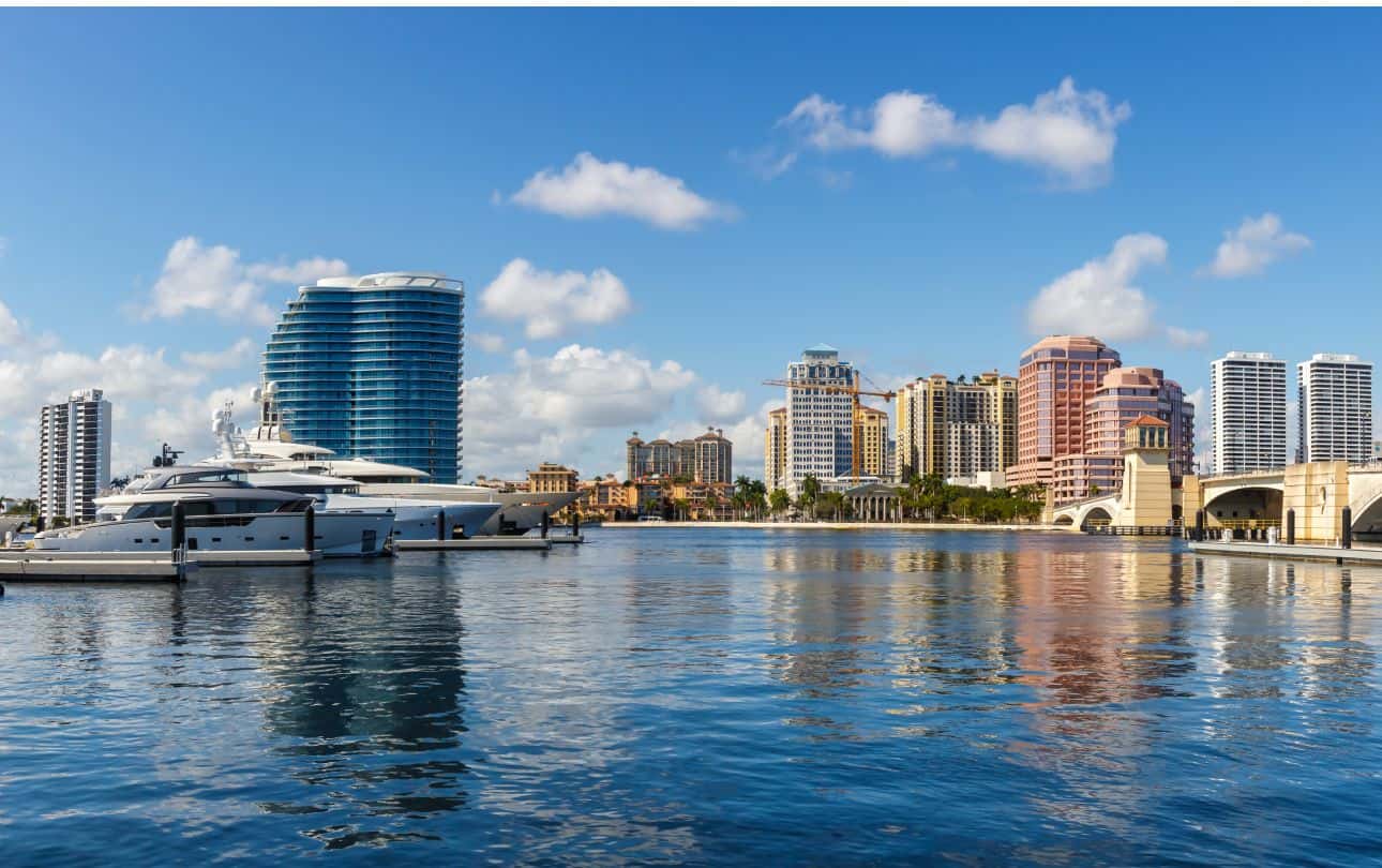 Waterfront cityscape with high-rise buildings, yachts, and a bridge under a blue sky.