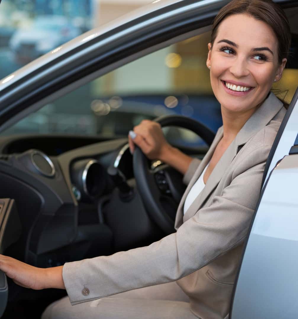 Smiling woman sitting in the driver’s seat of a vehicle at a dealership.