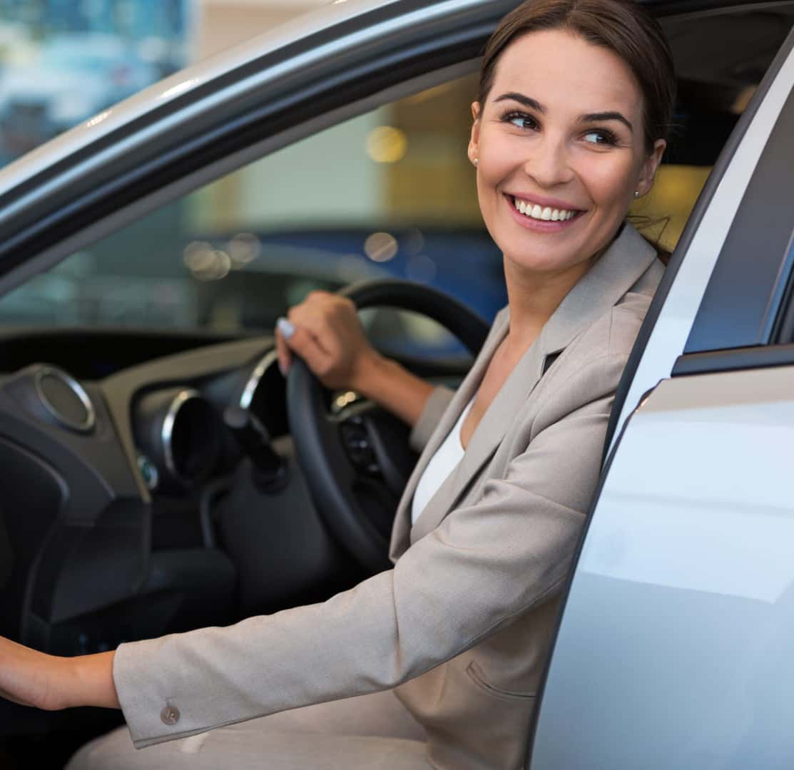 Woman smiling while seated in the driver’s seat of a vehicle at a dealership.