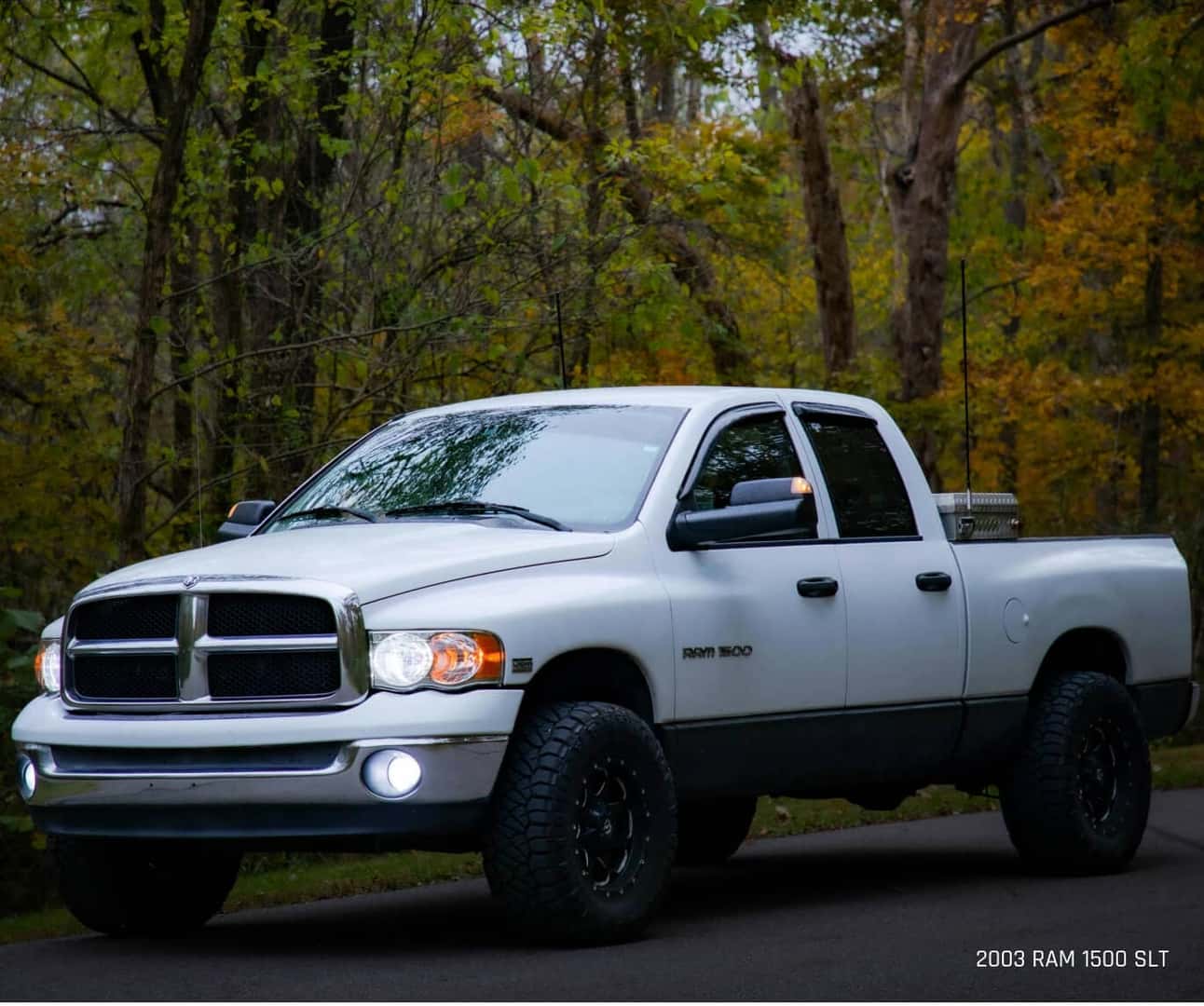 Three-quarter view of a Ram 1500 SLT in White parked on asphalt against a forest.