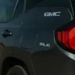 Close-up of the rear side of a dark-colored GMC vehicle, showing the "GMC" and "SLE" badges and a red tail light. The image emphasizes the vehicle's branding and trim level.