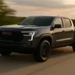 A black GMC pickup truck drives along a highway at sunset, with soft golden light highlighting its bold front grille and modern LED headlights. The motion blur in the background adds a dynamic sense of speed and performance.