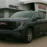 A gray GMC pickup truck is parked in front of a GMC dealership building, with prominent red and black GMC signage overhead. The dealership setting emphasizes new inventory and buying opportunities directly from the brand.
