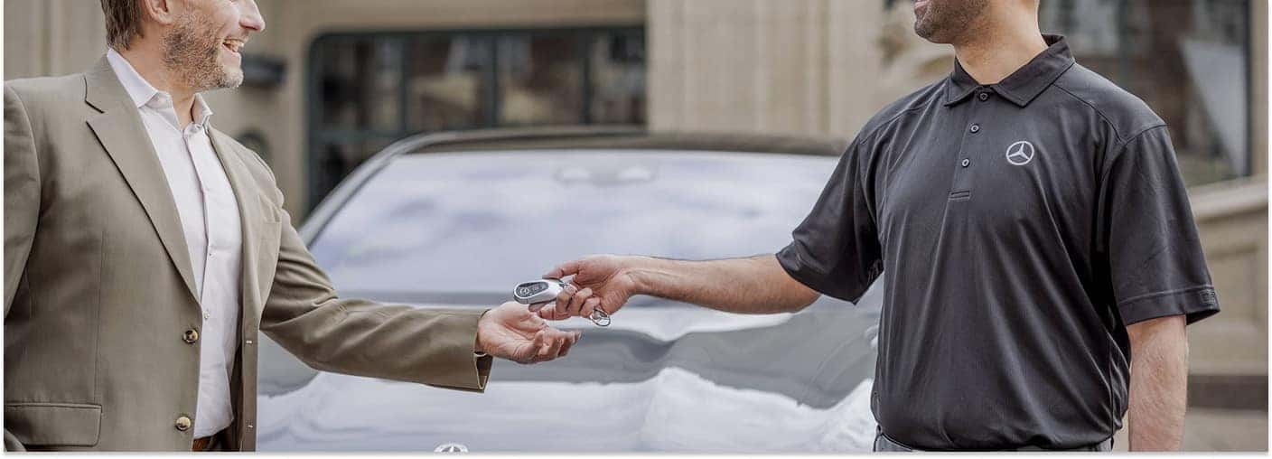 > A Mercedes-Benz representative in a branded black polo shirt hands a key fob to a smiling man in a suit jacket, with a blurred vehicle visible in the background.
