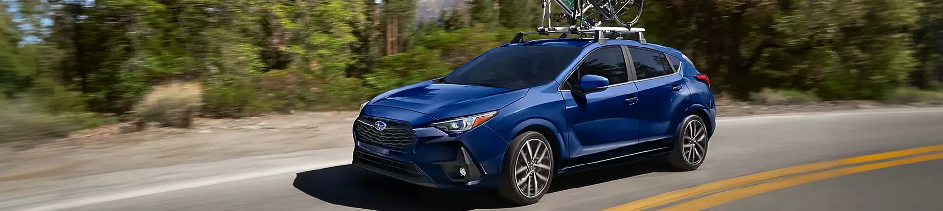 Blue 2026 Subaru Impreza carrying a bike on a roof rack along a tree-lined road