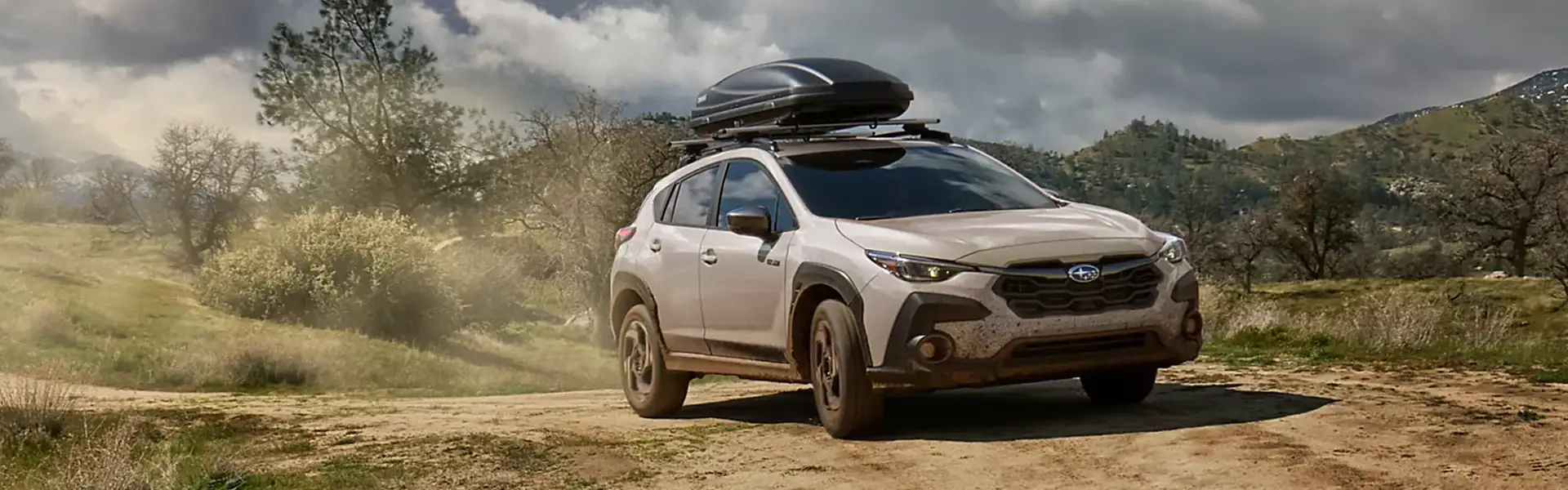 Beige Subaru Crosstrek with a rooftop cargo box on a dirt trail in the hills under a cloudy sky