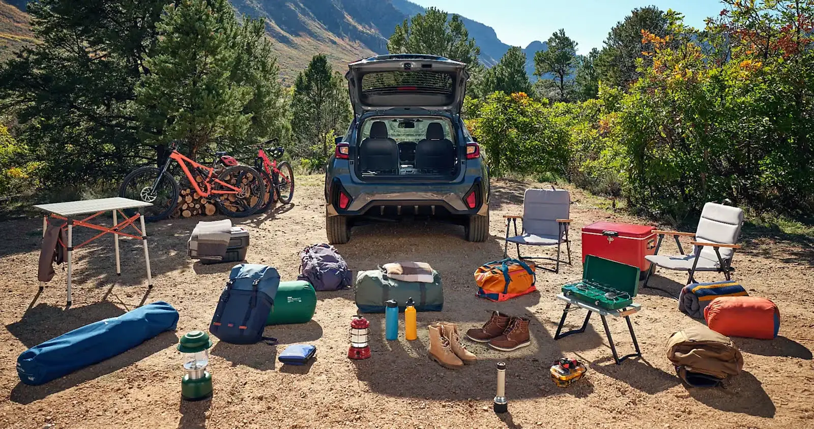 A 2026 Subaru Crosstrek with its tailgate open and a full kit of camping gear, bikes, coolers, and chairs laid out behind it at a desert campsite, the kind of investment routine maintenance protects