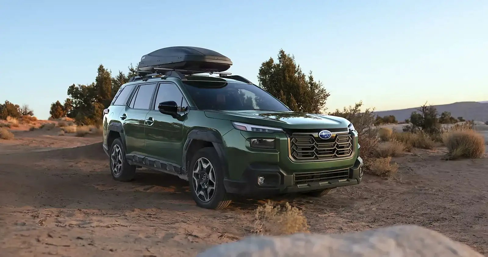 A green 2026 Subaru Outback Wilderness with a roof cargo box parked on desert terrain, the turbo and Wilderness trims that run tighter spark-plug and oil intervals in Las Vegas heat