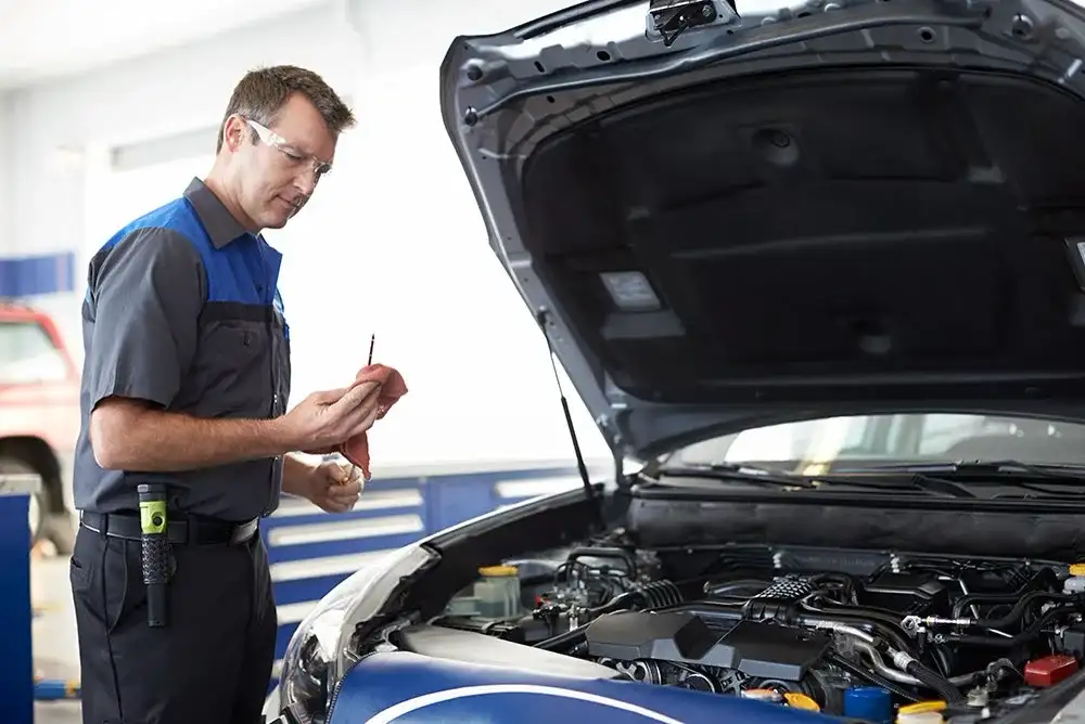 A Subaru-trained technician in safety glasses reading the oil dipstick at the open hood of a Subaru in the service bay