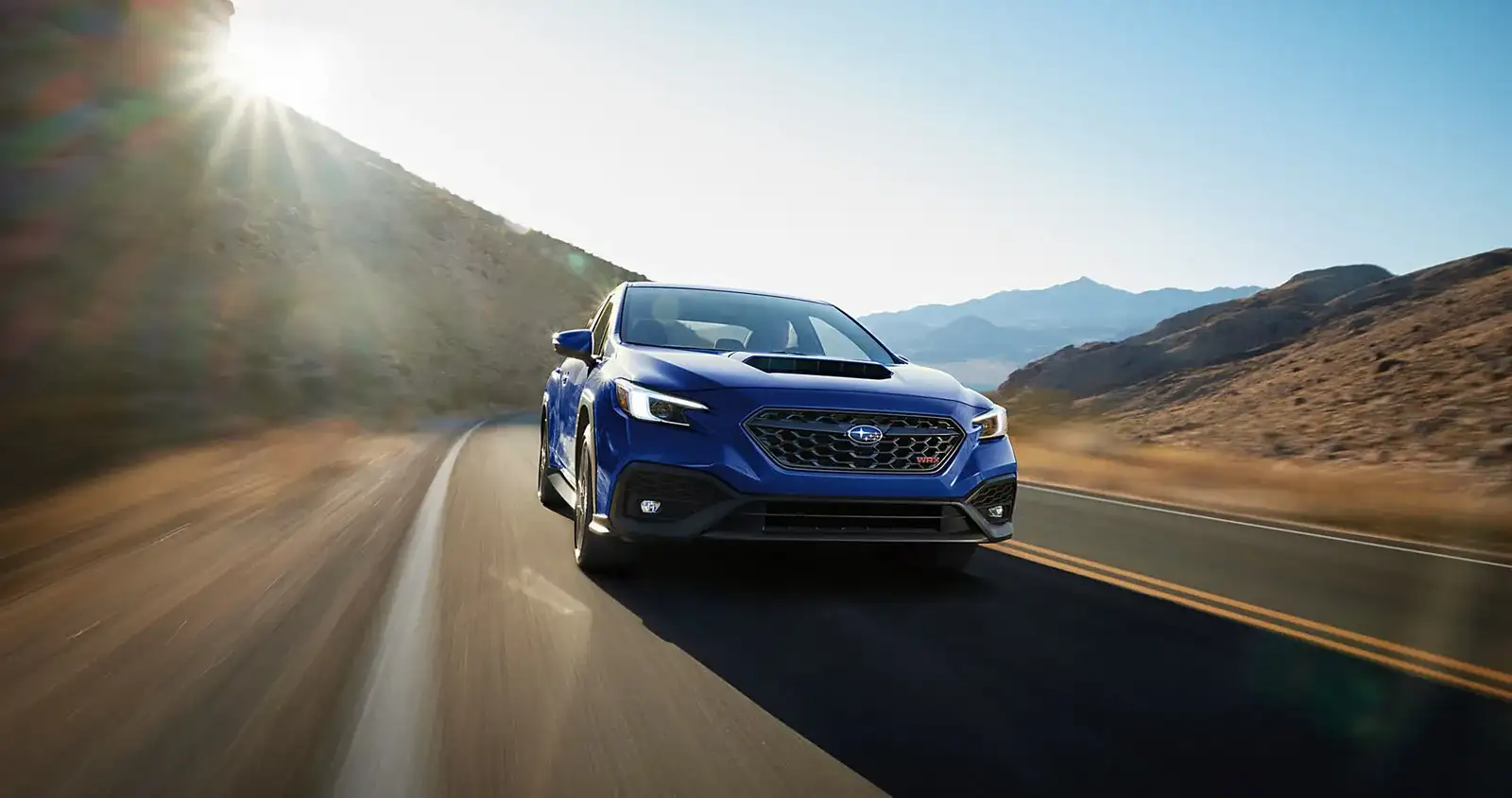A blue 2026 Subaru WRX in motion on a desert mountain road at golden hour, the performance model that runs the shortest oil, brake-fluid, and spark-plug intervals in the lineup