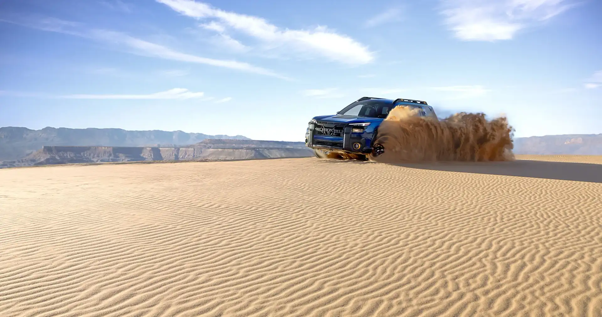 A blue 2026 Subaru Outback driving across desert sand and throwing up a rooster tail, demonstrating its standard Symmetrical all-wheel drive on loose terrain