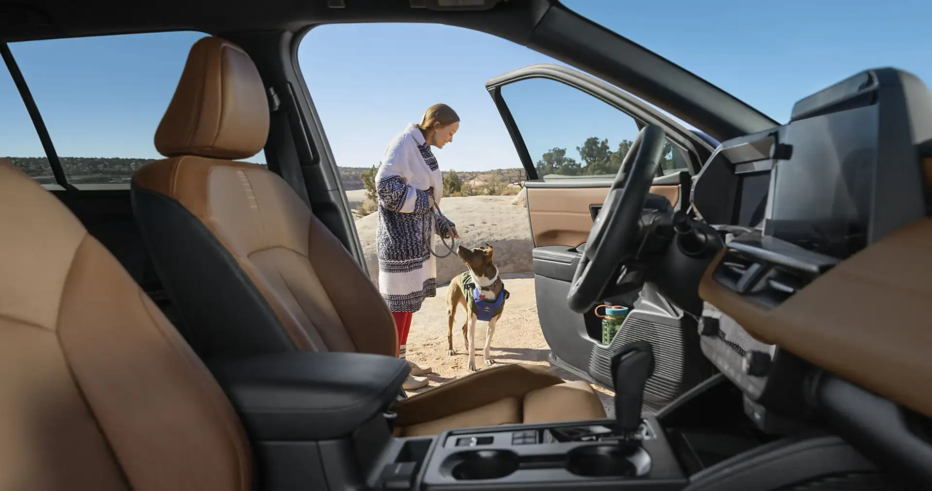 An owner standing with a dog beside the open door of a 2026 Subaru Outback at a desert trailhead, showing the brown leather interior and the large vertical touchscreen
