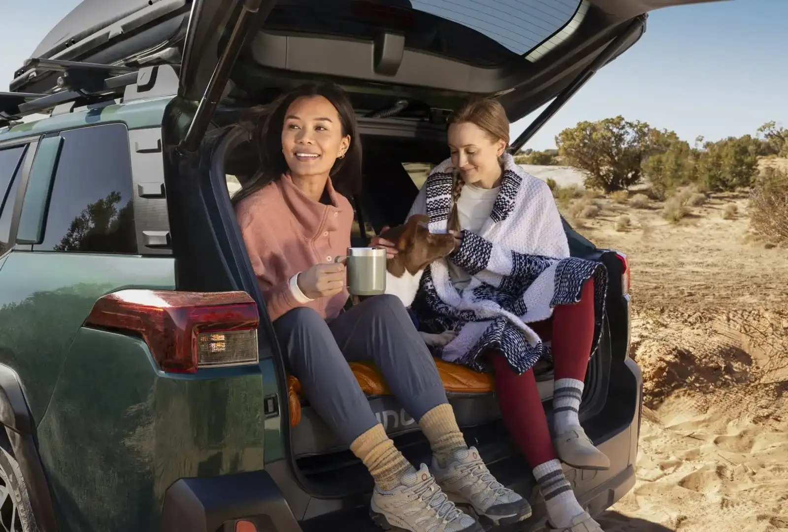 Two people sitting in the open rear cargo area of a 2026 Subaru Outback at a desert trailhead, wrapped in blankets with coffee