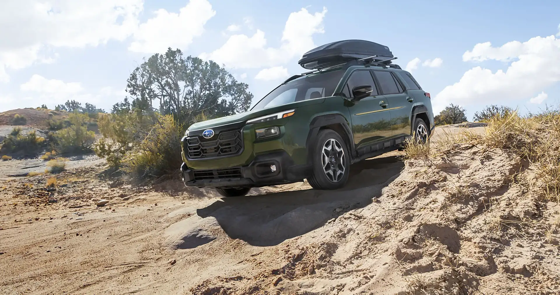 A green 2026 Subaru Outback Wilderness with a roof cargo box climbing a sandy desert grade, showing its higher ground clearance and standard all-wheel drive