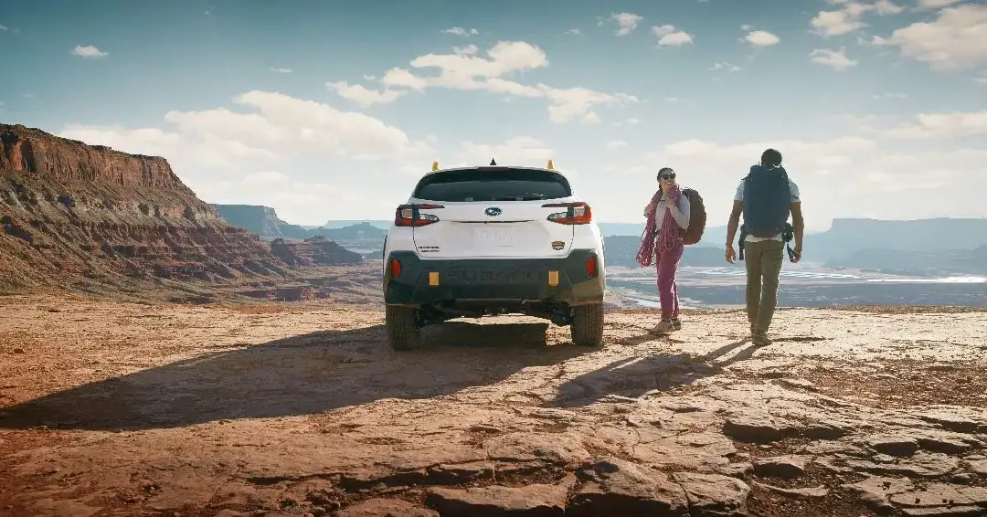 A white Subaru Crosstrek Wilderness parked on a slickrock mesa with two hikers walking toward the cliff edge with backpacks — the kind of sustained-heat trip that ages CVT fluid faster