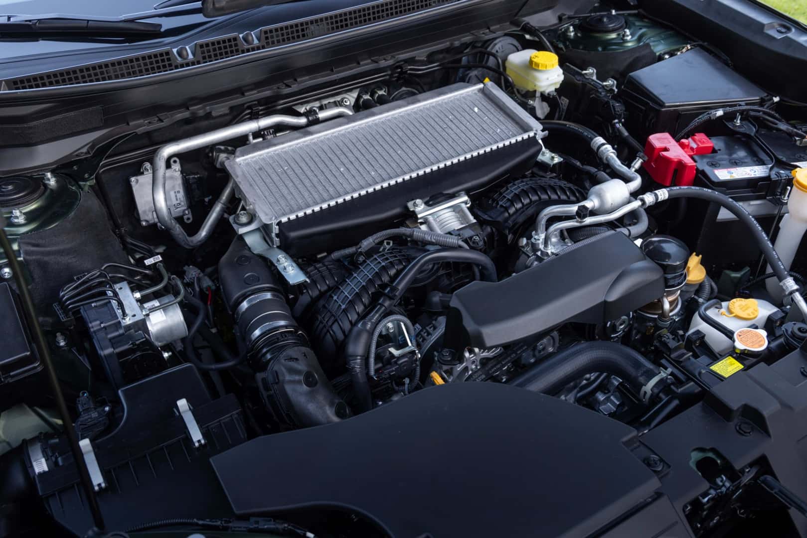 A Subaru turbo Boxer engine bay — intercooler on top, intake hose, brake-fluid reservoir with yellow OEM cap, and red battery terminal cap visible