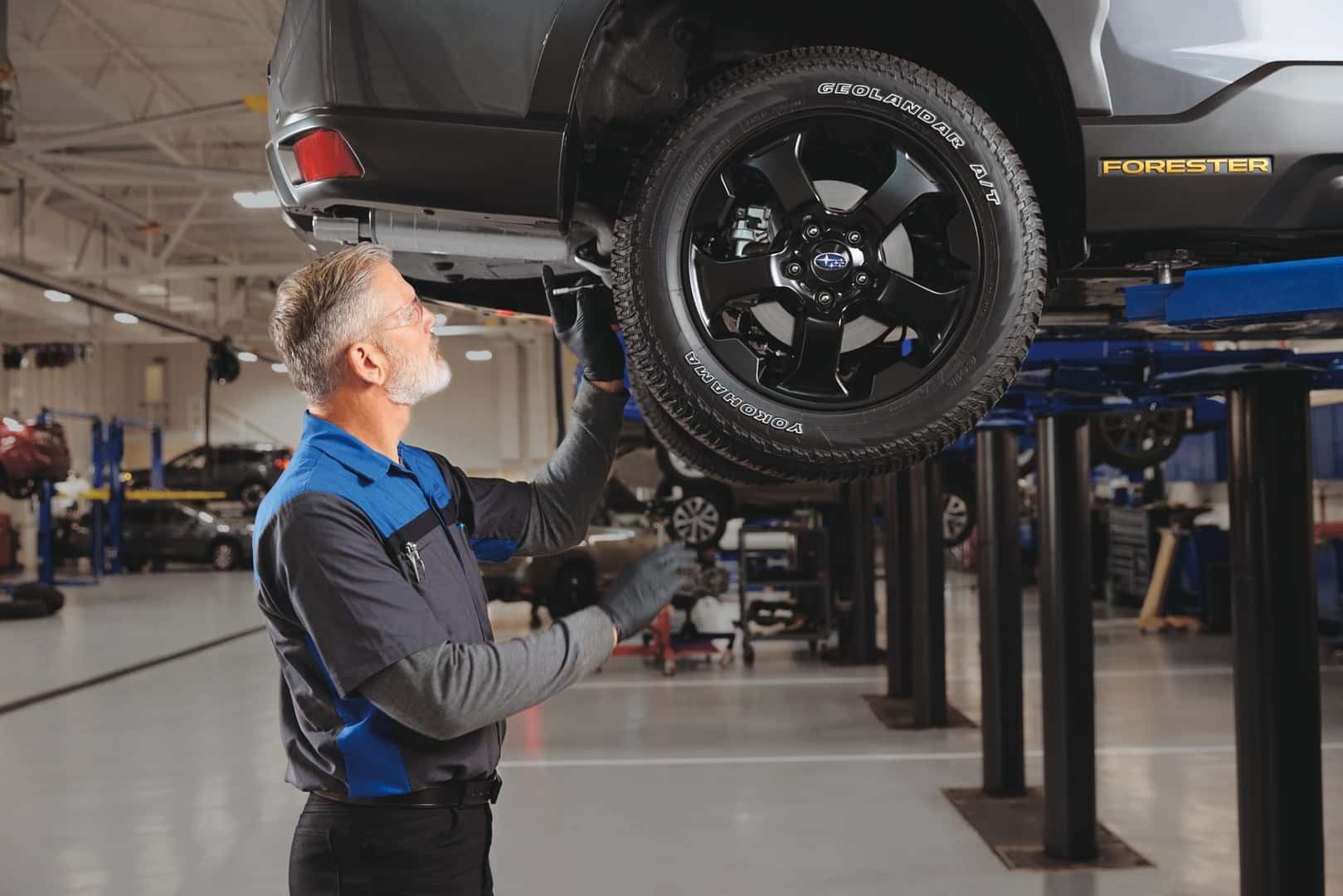 A senior Subaru technician in a Subaru-branded uniform inspecting a lifted gray Subaru Forester on a service-bay lift
