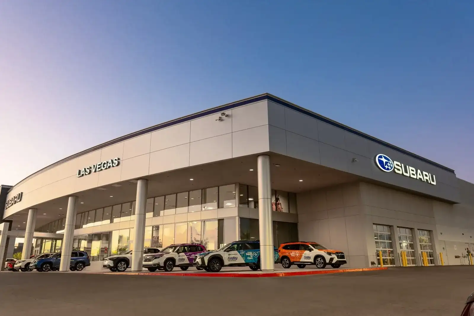 The Findlay Subaru of Las Vegas showroom at dusk, with the LAS VEGAS storefront sign and a row of Subaru Love Promise vehicles out front