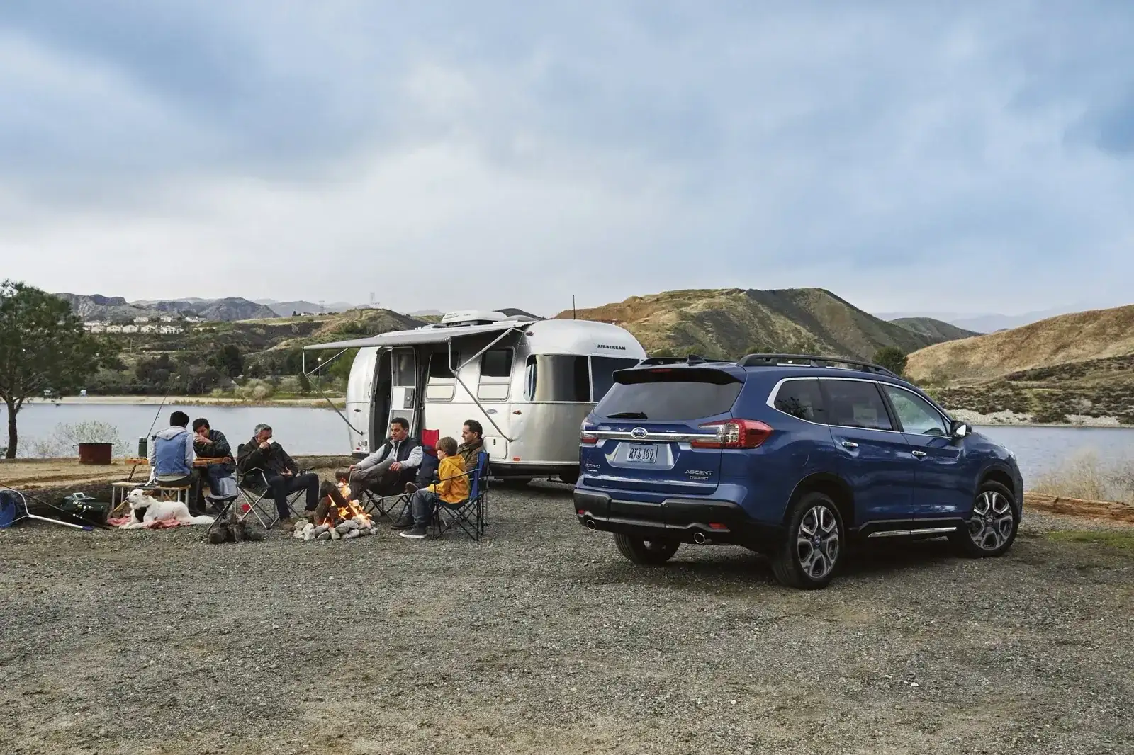 A Subaru Ascent towing an Airstream trailer at a lakeside campsite while a family gathers around a campfire