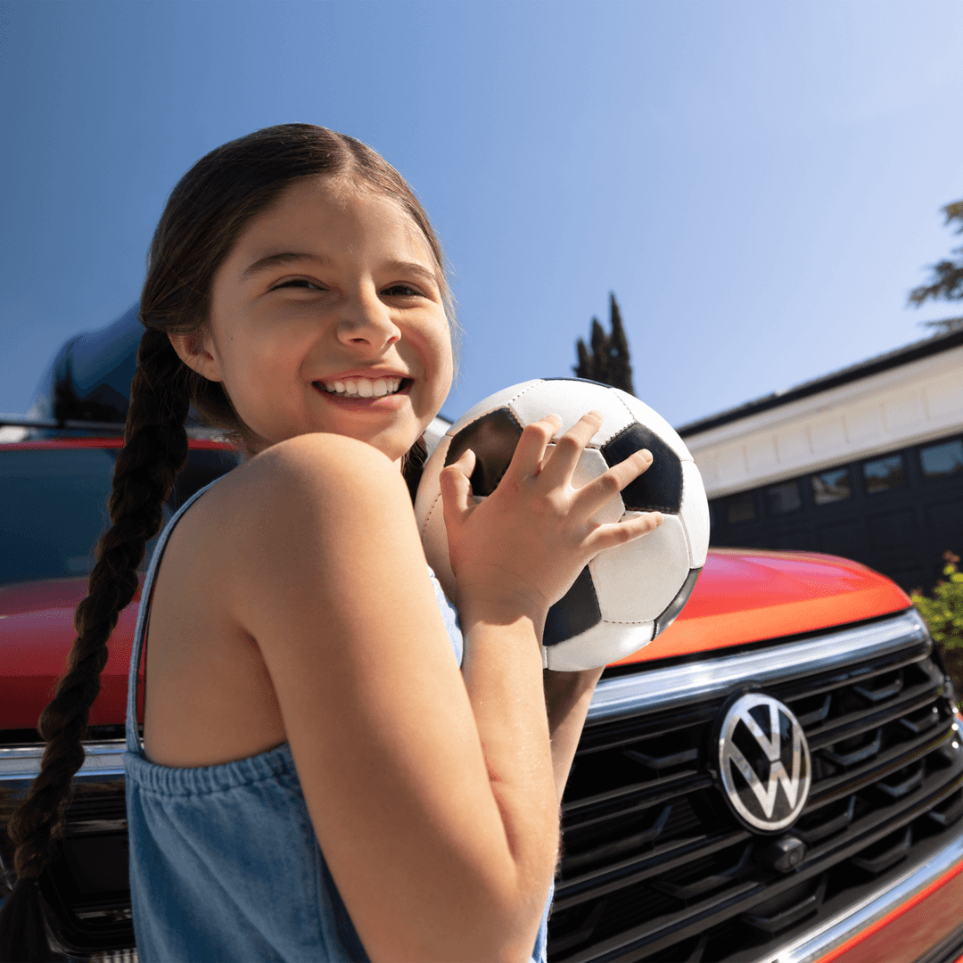 Smiling young girl holding a soccer ball in front of a red Volkswagen SUV, representing family fun in Twin Falls, Idaho