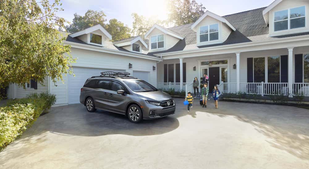 A gray Honda Odyssey minivan with a roof rack parked in a driveway as a family walks out of a house