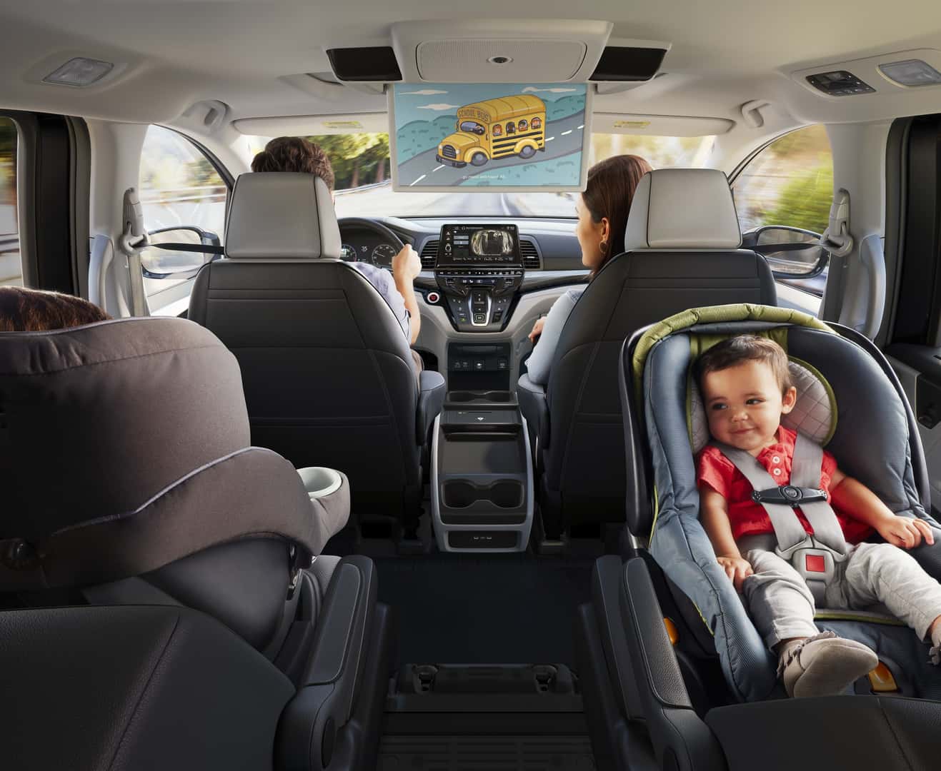 View from the back of a Honda Odyssey toward the front seats, with a ceiling-mounted rear entertainment screen, the center touchscreen, and two child car seats