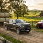 A gray Ram truck towing a trailer of hay bales alongside a red Ram truck on a farm path.