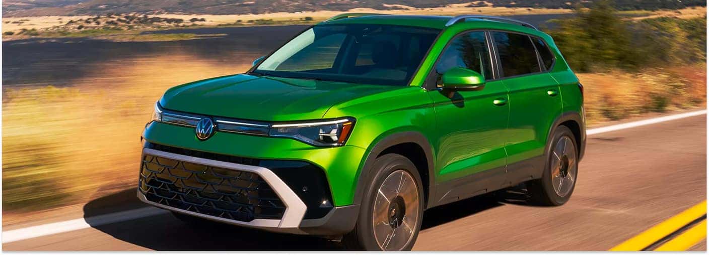 A green 2026 Volkswagen Taos SUV drives along an open asphalt road, showcasing its front profile against a backdrop of sunlit hills.