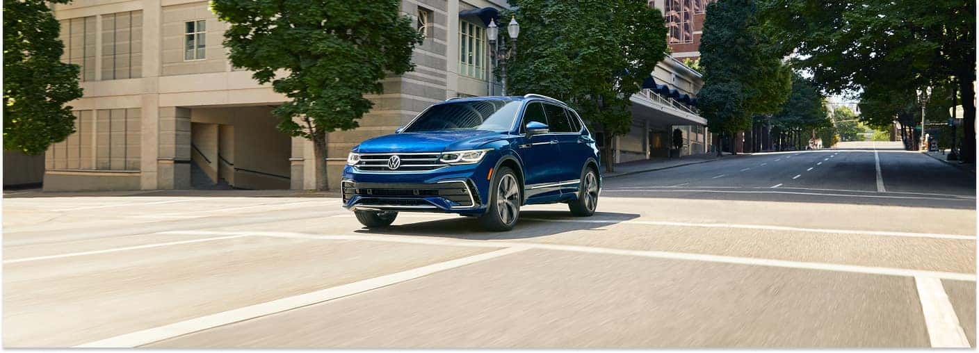 A blue 2026 Volkswagen Tiguan SUV drives through a city intersection, set against a backdrop of urban buildings and tree-lined streets.
