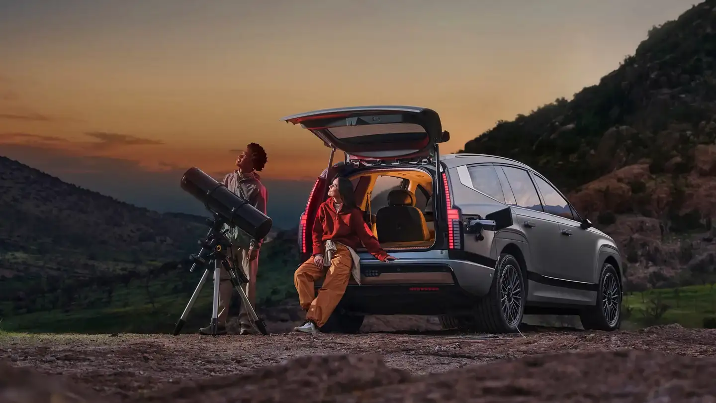 Hyundai IONIQ 9 electric SUV parked at a scenic overlook at sunset, with two people using a telescope beside the open rear hatch, showcasing cargo space and outdoor lifestyle.