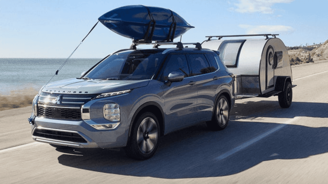 2024 Mitsubishi Outlander GT in blue with a roof cargo box and a compact trailer, driving along a coastal road.