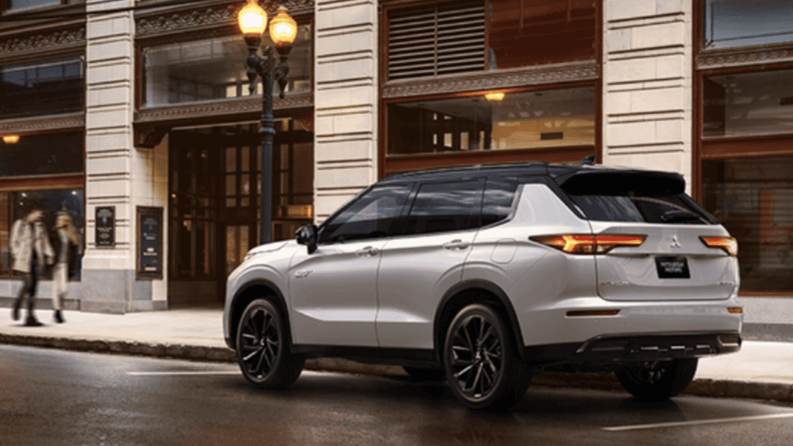 A silver 2025 Mitsubishi Outlander parked on a street in a city with a building facade and street lamps in the background.