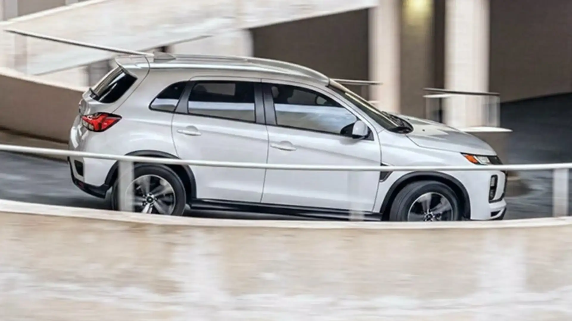 2026 Mitsubishi Outlander Sport white SUV driving beside a modern concrete building in a side profile view.