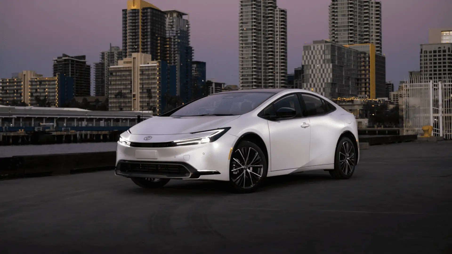 White 2025 Toyota Prius parked near an urban waterfront, highlighting aerodynamic exterior styling and efficient hybrid design in Hagerstown, MD.